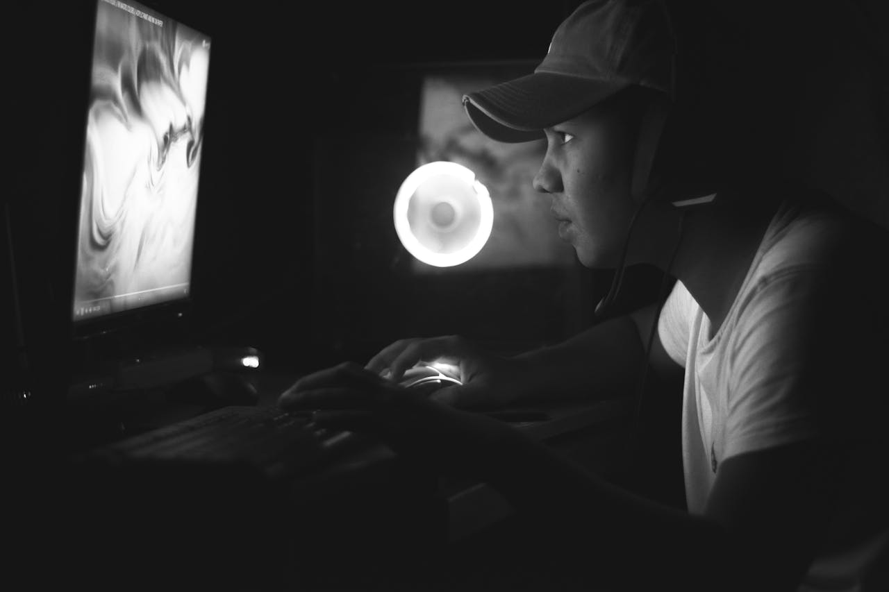 Side view of a gamer wearing a cap and headphones, illuminated by a computer screen in a dark room.