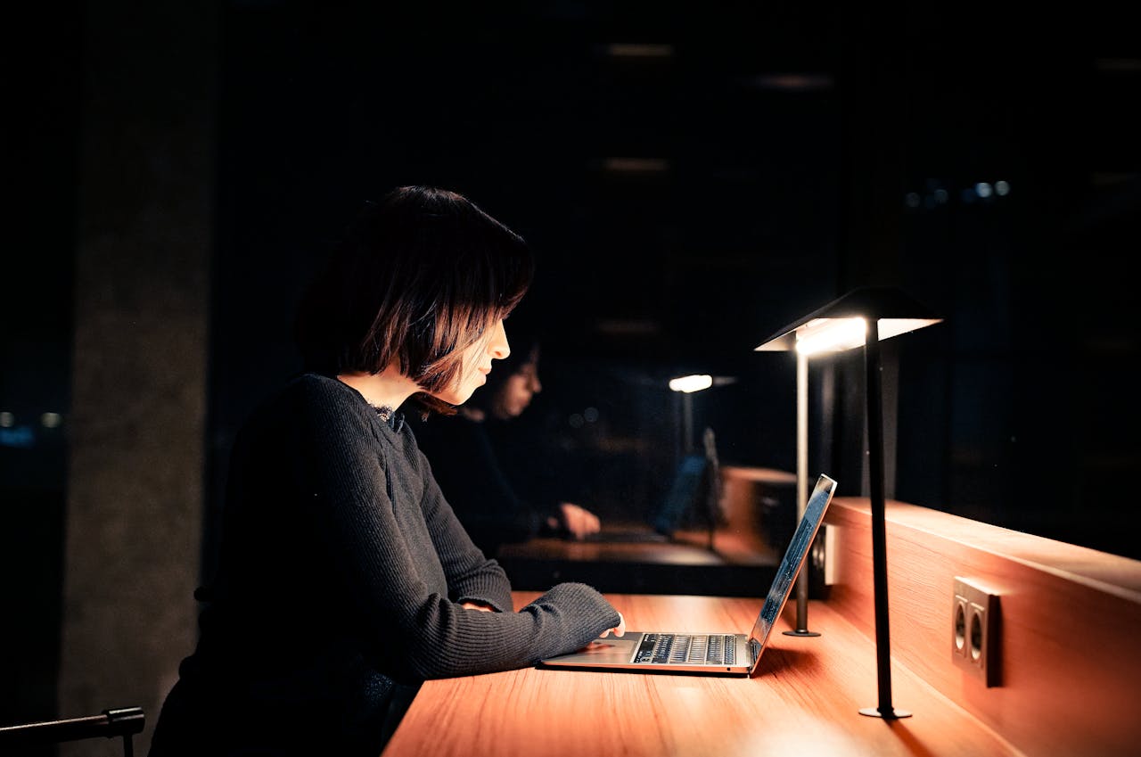 A woman focused on her laptop at a desk with dim lighting, working late at night.